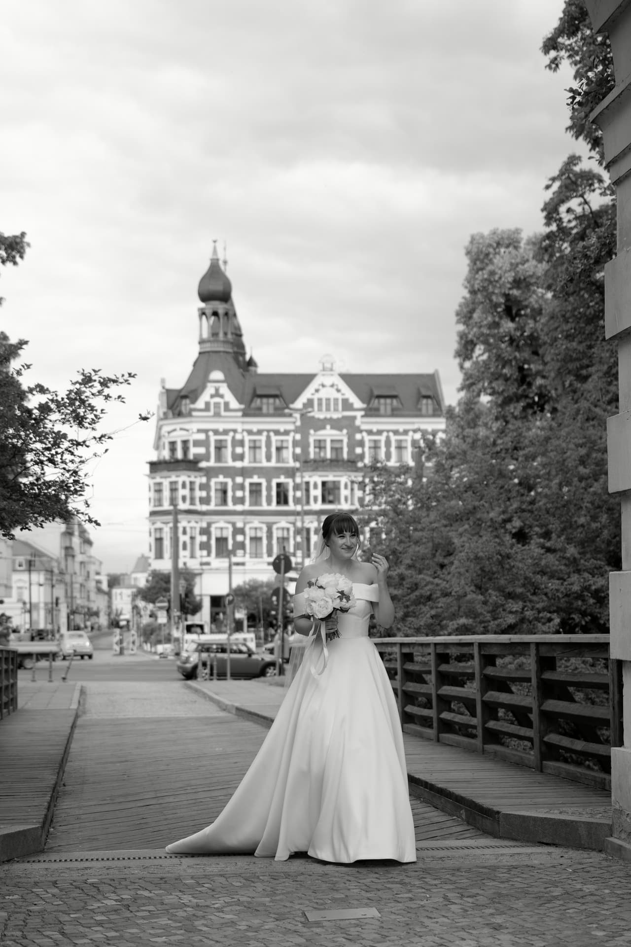 Schwarz-Weiß-Foto: Braut im Hochzeitskleid auf einer Holzbrücke vor einem historischen Gebäude