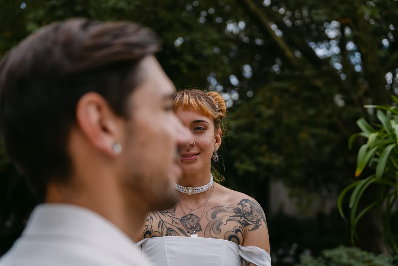 Braut mit Tattoos und Perlenhalsband lächelt hinter dem Bräutigam – ungestellte Hochzeitsfotografie