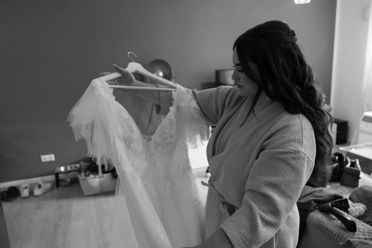 Schwarz-Weiß-Foto: Braut beim Getting Ready mit ihrem Hochzeitskleid am Bügel