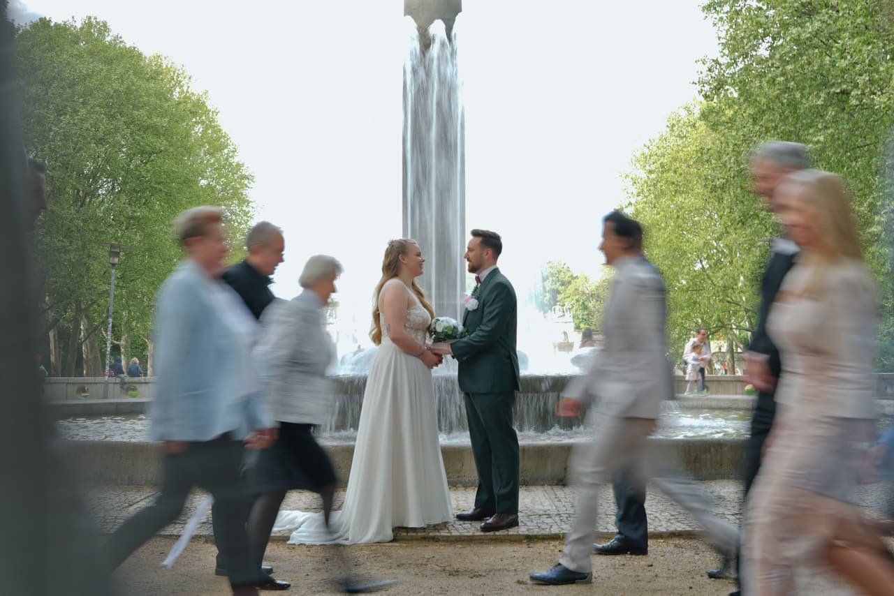 Brautpaar steht ruhig am Springbrunnen, während die Hochzeitsgäste unscharf vorbeigehen