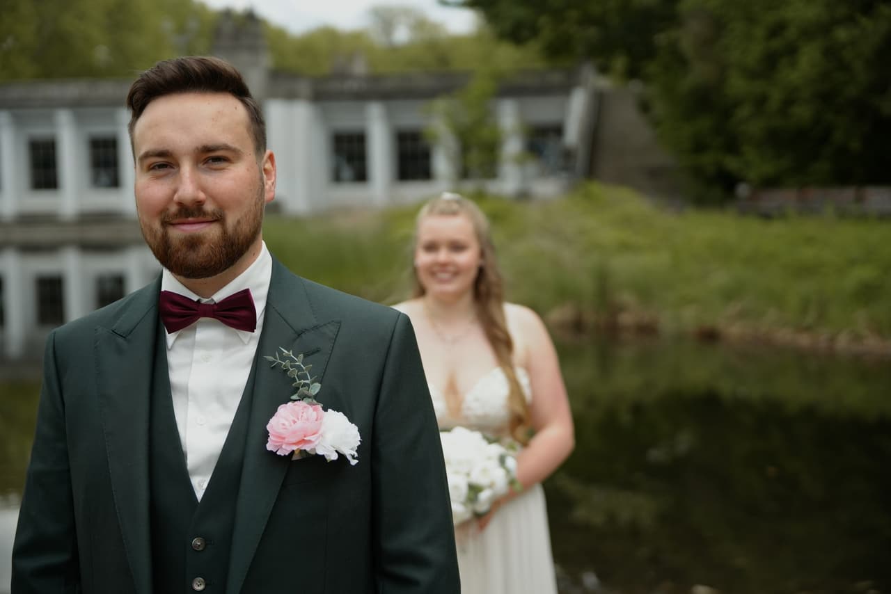 Portrait des Bräutigams mit Fliege und Ansteckblume, die Braut wartet im Hintergrund