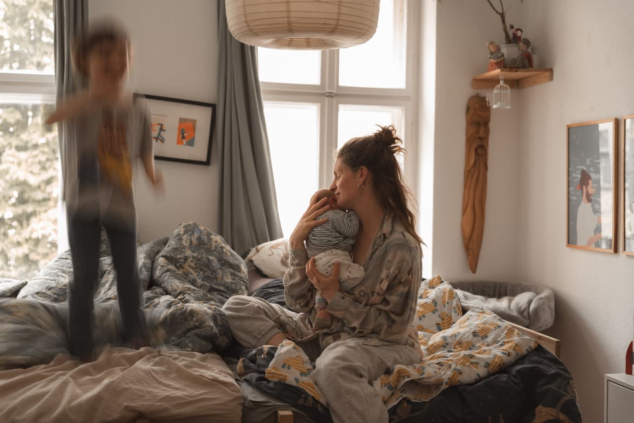 Mutter mit Neugeborenem auf dem Bett, das Geschwisterkind springt im Hintergrund